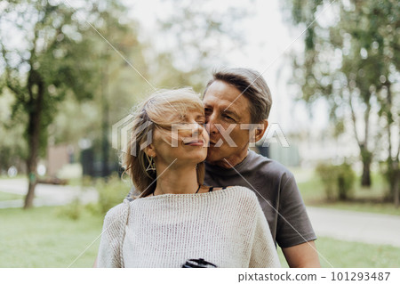 An adult mature happy couple in love hugging outdoors in city park. A blonde caucasian man and woman spend time together and drinking coffee. Senior wife and husband walking outside. 101293487