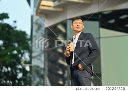 Confident young businessman in elegant suit holding paper cup of coffee standing outdoors in business district 101294320