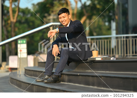 Full length of young businessman sitting on stairs outside office building and checking time on his wristwatch 101294325