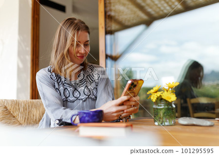 Young woman in ethnic outfit sitting at table with phone, working, having mug of morning coffee at home. Young woman in ethnic outfit sitting at table with phone, working, having mug of morning coffee at home. 101295595