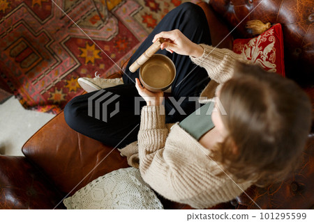 Young pretty caucasian girl playing on chakra cleaning tibetan singing bowl, while sitting on sofa at home interior. Yoga concept, meditation and sound therapy. Lifestyle concept. Top view 101295599