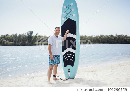 Joyful smiling male surfer on the beach. Young man in light t-shirt and multi colored shorts holding paddleboard. Summertime sport and fit concept. Joyful smiling male surfer on the beach. Young man in light t-shirt and multi colored shorts holding paddleboard. Summertime sport and fit concept. 101296759