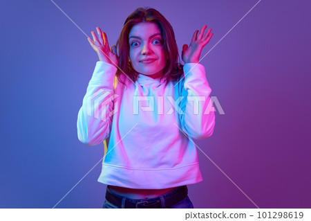 Emotional young girl with big eyes looking at camera with questioning face, posing against gradient blue purple studio background in neon light 101298619