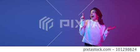 Excited, happy, positive young girl pointing with finger up against gradient blue purple studio background in neon light. Banner 101298624