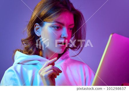 Close-up image of beautiful young girl looking on tablet with interest and excitement, reading news against purple background in neon light 101298637