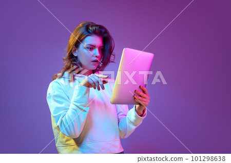Astonished, surprised. Young pretty girl looking on tablet with shocked and excited face against purple background in neon light 101298638