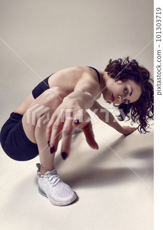 Beautiful, confident young woman with curly hair posing in sportswear and sneakers against light grey studio background. 101303719