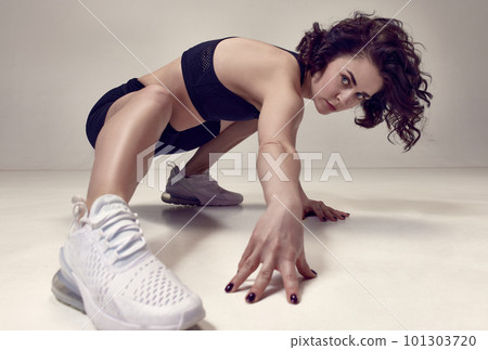 Beautiful, confident young woman with curly hair posing in sportswear and sneakers against light grey studio background. 101303720