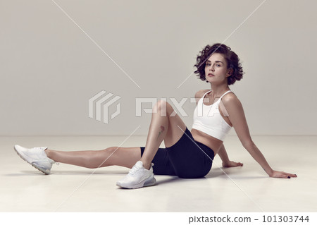 Young beautiful woman with curly short hair, in comfortable sportswear sitting on floor after workout against grey studio background 101303744