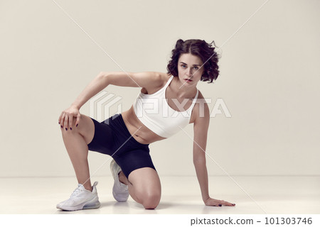 Pretty young woman with curly hair and fit, muscular, slim body in sportswear posing against light beige studio background 101303746