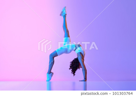 Flexible, sportive young woman training, doing stretching exercises against gradient pink blue background in neon light 101303764