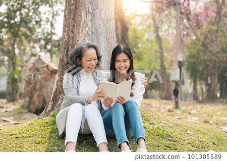Lovely granddaughter and grandmother sitting under the tree in the green park, read a book together Lovely granddaughter and grandmother sitting under the tree in the green park, read a book together 101305589