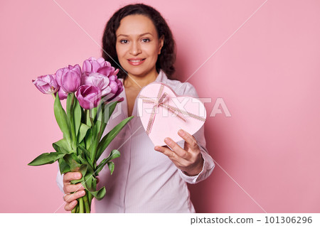 Details on a bouquet of purple tulips and a cute heart shaped gift box in the hands of blurred happy smiling woman, isolated over pink background. Happy Mother's Day. International Women's Day concept Details on a bouquet of purple tulips and a cute heart shaped gift box in the hands of blurred happy smiling woman, isolated over pink background. Happy Mother's Day. International Women's Day concept 101306296