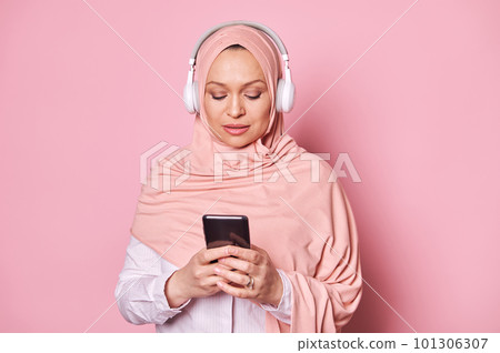 Close-up portrait of a Middle-Eastern Muslim woman in pink hijab, using mobile phone, scrolling playlist, checking mobile apps, enjoying listening to music in headphones, isolated over pink background Close-up portrait of a Middle-Eastern Muslim woman in pink hijab, using mobile phone, scrolling playlist, checking mobile apps, enjoying listening to music in headphones, isolated over pink background 101306307