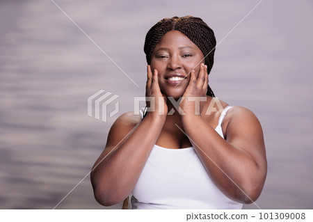 Cheerful woman using face moisturizing cream for skincare routine and looking at camera. African american body positive lady doing spa beauty facial procedures and posing for cosmetology commercial Cheerful woman using face moisturizing cream for skincare routine and looking at camera. African american body positive lady doing spa beauty facial procedures and posing for cosmetology commercial 101309008