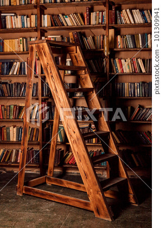 A large wooden staircase stands in front of a shelf with books in a dark room A large wooden staircase stands in front of a shelf with books in a dark room 101309941