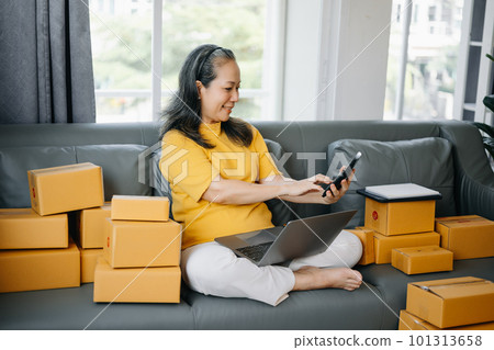 Startup small business SME, Entrepreneur owner Senior woman using smartphone or tablet taking receive and checking online purchase shopping order to preparing pack product box. 101313658