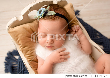 A cute newborn baby girl in a white jumpsuit with a flower bandage sleeps in a wooden crib on a brown pillow, top view. On a light wooden background.  101314176