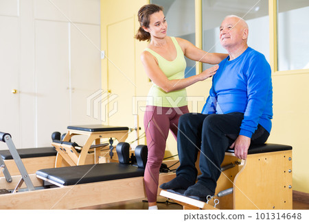 Elderly man practicing Pilates system on reformer supervised by qualified trainer Elderly man practicing Pilates system on reformer supervised by qualified trainer 101314648