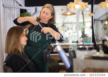 Aged woman hair stylist making haircut to young female client 101315344