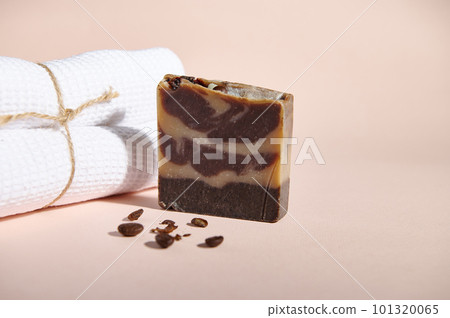 Still life with a bar of organic homemade cold pressed soap or solid body scrub, with natural ingredients and ground coffee beans next to a white bath towel on pink backdrop. Anti-cellulite body care 101320065
