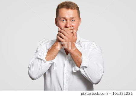 Handsome young male with blue eyes and healthy skin, covers mouth with both palms, tries to be speechless, doesnt want to reveal secret, dressed in elegant white shirt in one tone with background 101321221