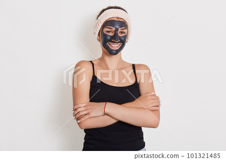 Close up portrait of young female applies homemade facial clay mask, has white hairband around head, smiles happily, keeps hands folded against white background. 101321485