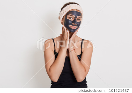 Pleased european woman applies nourishing clay mask on face, has glad expression, looks directly at camera, touches cheeks, having problem of dry skin. Pleased european woman applies nourishing clay mask on face, has glad expression, looks directly at camera, touches cheeks, having problem of dry skin. 101321487