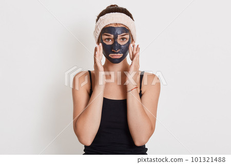 Young unhappy housewife with black cosmetic mask on face standing isolated over white background, touching her cheeks with fingers, looking at camera. 101321488