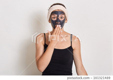 Pretty girl with black clay facial mask, surprised girl with hair band on her head, having astonished facial expression, posing isolated over background. Skin care, beauty treatment. Pretty girl with black clay facial mask, surprised girl with hair band on her head, having astonished facial expression, posing isolated over background. Skin care, beauty treatment. 101321489