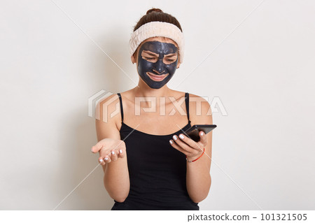 Young lady with black facial cosmetic mask using cellphone, looking directly at device screen, spreading palm, having astonished and puzzled facial expression. Young lady with black facial cosmetic mask using cellphone, looking directly at device screen, spreading palm, having astonished and puzzled facial expression. 101321505