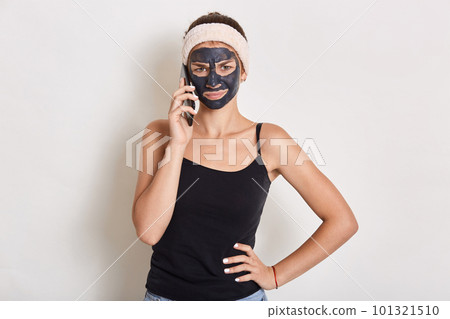 Young teen girl with black clay facial mask making phone call, standing isolated over white background, wearing headband and t shirt, keeping hands on hip, looks at camera. Young teen girl with black clay facial mask making phone call, standing isolated over white background, wearing headband and t shirt, keeping hands on hip, looks at camera. 101321510