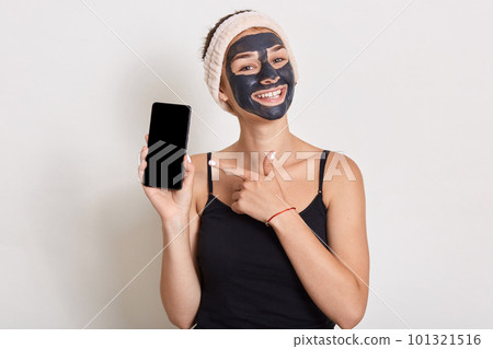 Charming lady with cosmetic facial mask holding modern mobile phone in hands and pointing with index finger at copy spy space against white wall, happy lady doing beauty procedures. Charming lady with cosmetic facial mask holding modern mobile phone in hands and pointing with index finger at copy spy space against white wall, happy lady doing beauty procedures. 101321516