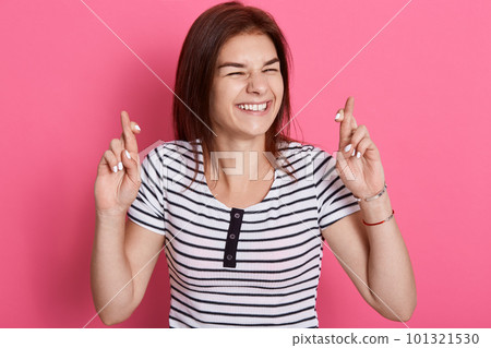Hopeful dark hared woman feels lucky, crosses fingers, awaits for miracle, makes wish and has high hopes, smiles broadly, wears striped t shirt, poses against rose wall. Hopeful dark hared woman feels lucky, crosses fingers, awaits for miracle, makes wish and has high hopes, smiles broadly, wears striped t shirt, poses against rose wall. 101321530
