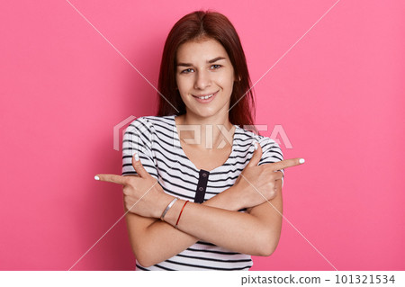 Cheerful dark haired woman crosses arms over chest, points left and right, gives two variants, advertises products, female stands against pink background. Cheerful dark haired woman crosses arms over chest, points left and right, gives two variants, advertises products, female stands against pink background. 101321534