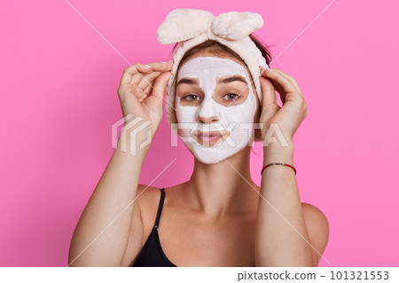Young lady wearing sleeveless t shirt and headband, looking at camera with cosmetic mask on face, doing beauty procedures in morning at home. 101321553