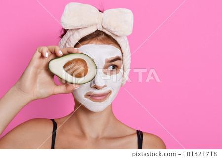 Young woman holding avocado in hands and covering her eyes with fruit, having white mask on face, looking aside, wearing head band with bow isolated over rose background. Young woman holding avocado in hands and covering her eyes with fruit, having white mask on face, looking aside, wearing head band with bow isolated over rose background. 101321718