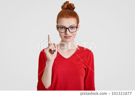 Clever freckled foxy student gets brilliant idea in mind, raises index finger in eureka gesture, wears transparent glasses, red sweater, poses against white background, has red hair combed in bun Clever freckled foxy student gets brilliant idea in mind, raises index finger in eureka gesture, wears transparent glasses, red sweater, poses against white background, has red hair combed in bun 101321877