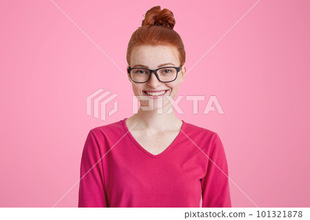 Isolated shot of happy ginger woman in pink sweater, wears square spectacles, has red hair tied in knot, freckled skin, pleasant smile, isolated over pink background. Cheerful female student indoor Isolated shot of happy ginger woman in pink sweater, wears square spectacles, has red hair tied in knot, freckled skin, pleasant smile, isolated over pink background. Cheerful female student indoor 101321878
