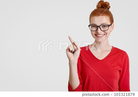 Intrigued ginger female teacher in spectacles attracts your attention to blank copy space, indicates with index finger, has charmig smile, likes students project work, poses over white background Intrigued ginger female teacher in spectacles attracts your attention to blank copy space, indicates with index finger, has charmig smile, likes students project work, poses over white background 101321879