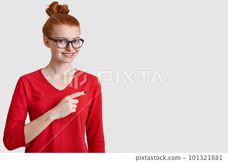 Studio shot of cheerful woman with red hair bun, wears spectacles, casual clothes, points aside with index finger, shows free space, isolated over white background, has freckled skin, gentle smile Studio shot of cheerful woman with red hair bun, wears spectacles, casual clothes, points aside with index finger, shows free space, isolated over white background, has freckled skin, gentle smile 101321881