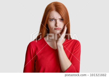 Displeased red haired woman with freckled skin, keeps hand on cheek, suffers from toothache, has sensitivity, wears casual red clothes, isolated over white background. Dental problems concept Displeased red haired woman with freckled skin, keeps hand on cheek, suffers from toothache, has sensitivity, wears casual red clothes, isolated over white background. Dental problems concept 101321882
