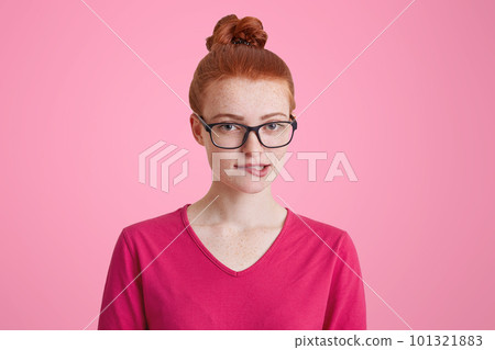 Freckled female student with ginger hair bun wears big square spectacles, bites lower lips as tries to recollect something in mind, dressed casually, isolated over pink background. Beauty concept Freckled female student with ginger hair bun wears big square spectacles, bites lower lips as tries to recollect something in mind, dressed casually, isolated over pink background. Beauty concept 101321883