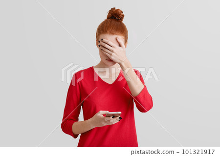 Portrait of beautiful freckled young woman covers face with hand, uses modern mobile phone for online communication, isolated over white studio background. Technology and people concept 101321917