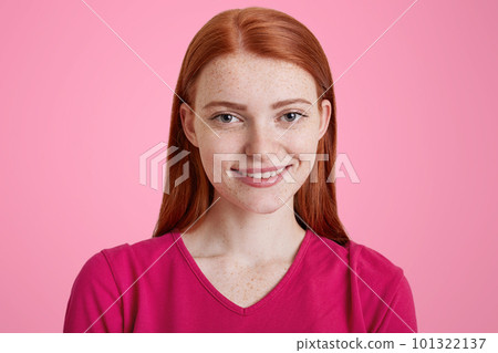 Beautiful ginger female with pleasant smile, has freckled skin, being glad to be photographed, dress in pink sweater, isolated over studio background. Red haired pretty young woman poses indoor Beautiful ginger female with pleasant smile, has freckled skin, being glad to be photographed, dress in pink sweater, isolated over studio background. Red haired pretty young woman poses indoor 101322137