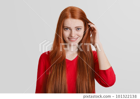 Portrait of redhead young female with long hair, has freckled face, pleasant smile, touches hair, isolated over white background. Ginger woman poses in studio, happy be photograhed. 101322138
