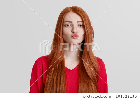 Adorable red haired female with freckled skin, rounds lips, going to kiss someone, has long redish hair, isolated over white background. Natural beautiful woman poses indoor. Body language concept Adorable red haired female with freckled skin, rounds lips, going to kiss someone, has long redish hair, isolated over white background. Natural beautiful woman poses indoor. Body language concept 101322139