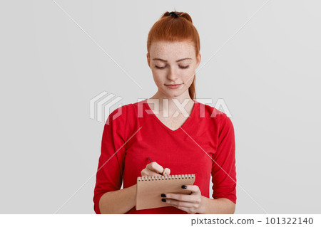 Serious freckled female in red sweater writes notes in spiral diary with pen. Beautiful ginger woman poses with notebook, isolated over white background. Student prepares for exams, writes information Serious freckled female in red sweater writes notes in spiral diary with pen. Beautiful ginger woman poses with notebook, isolated over white background. Student prepares for exams, writes information 101322140