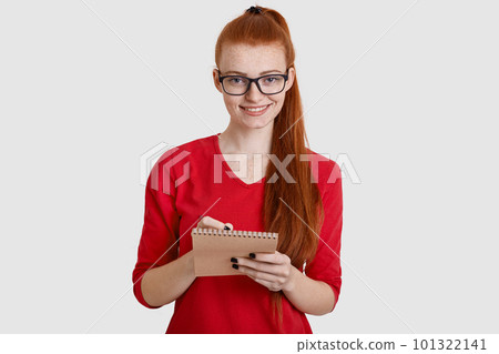 Freckled ginger pretty woman with pony tail, writes down in notepad with pen, records information, smiles gently, isolated over white background. Red haired student prepares for examination. Freckled ginger pretty woman with pony tail, writes down in notepad with pen, records information, smiles gently, isolated over white background. Red haired student prepares for examination. 101322141