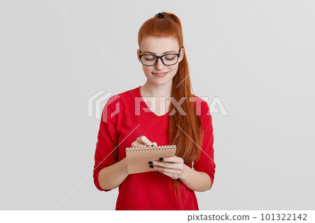Pretty young woman with long ginger ponytail, writes notes in notebook, creates her reminder or working schedule, writes down information, isolated over white background. Female student studies Pretty young woman with long ginger ponytail, writes notes in notebook, creates her reminder or working schedule, writes down information, isolated over white background. Female student studies 101322142